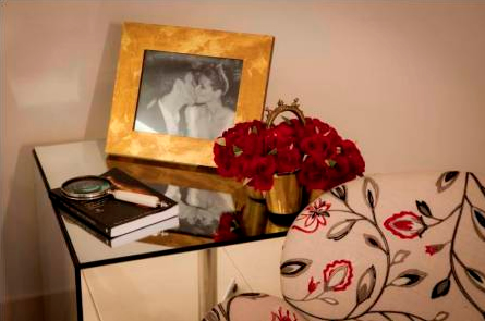 Bloco espelhado com porta retrato com a foto de um casal se beijando, vaso de flor vermelho ao lado e um sofá.