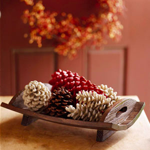 Arranjo de mesa composto por uma pequena bandeja de madeira com cinco pinhas, sendo uma marrom, três brancas e uma vermelha.