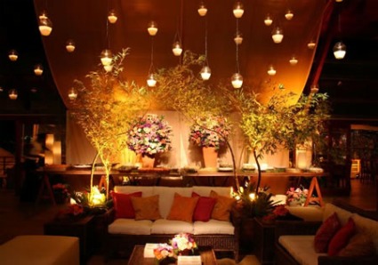 Ambiente com baixa iluminação, cheio de vasos de flores, duas árvores e sofás brancos com almofadas rosa e vermelhas.