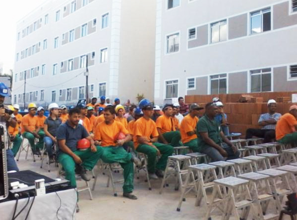 Grupo de trabalhadores em uma construção sentados em bancos pratas na área externa prestando atenção em um evento.