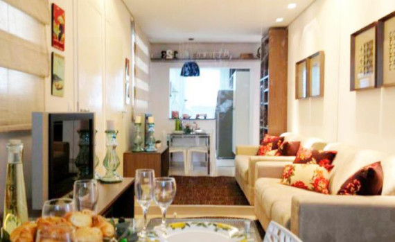 Sala de jantar com mesa, cadeiras, taças, sofá com almofadas, rack com televisão, tapete e espelho grande na parede.
