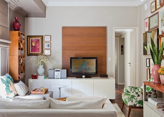 Sala de estar com sofá, almofadas, rack, televisão, aparelho de som, painel de madeira e quadros na parede.
