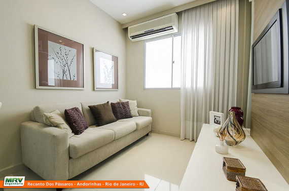 Sala de estar com sofá, almofadas, painel com televisão, rack com vasos e quadros, ar condicionado e cortina.