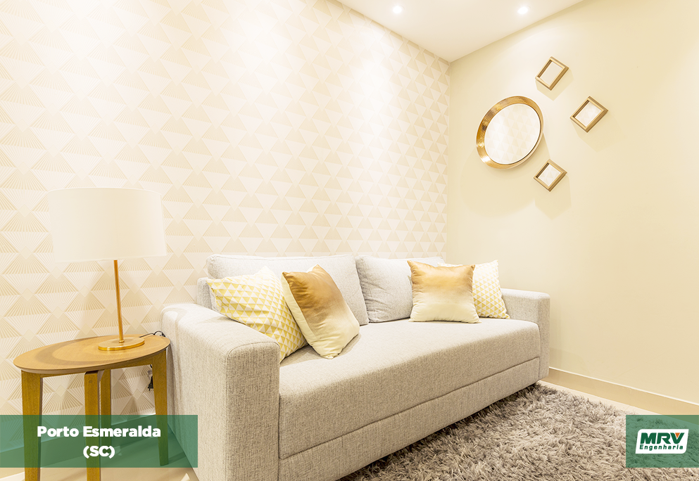 Sala com papel de parede em tons bege, sofá cinza com almofadas em tons de amarelo, mesa de canto e decorações de parede