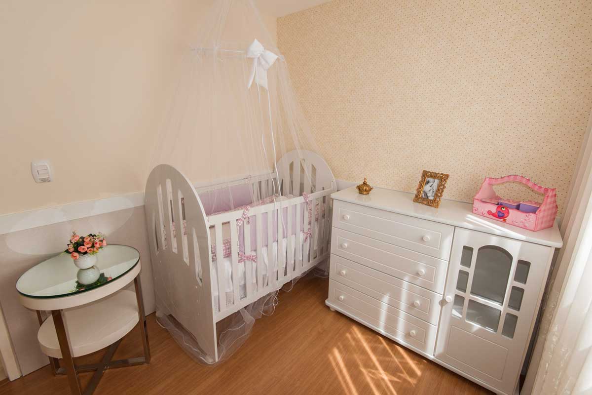Quarto de bebê com berço branco, mosqueteiro, cômoda branca com quatro gavetas e mesa espelhada ao lado.