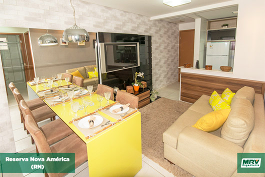 Sala de estar conjugada com a de jantar, decoração em amarelo, sofá, almofada, mesa com quatro cadeiras painel e televisão.