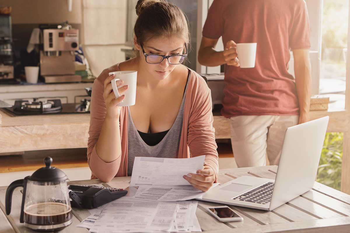 Mulher tomando café e vendo contas na cozinha. Em frente um notebook e celular. Atrás um homem segurando uma xícara.