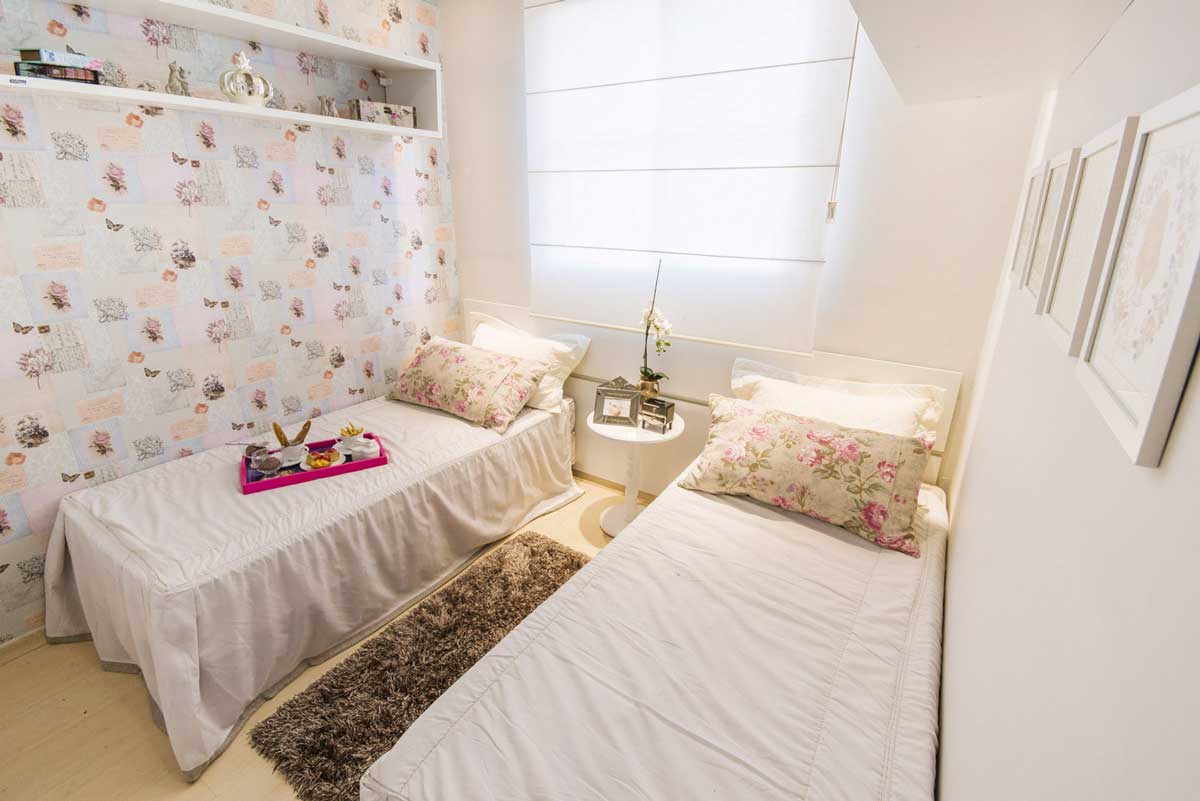 Quarto de solteiro com duas camas, lençois, travesseiros, mesa de centro embaixo da janela, cortina e parede florida.